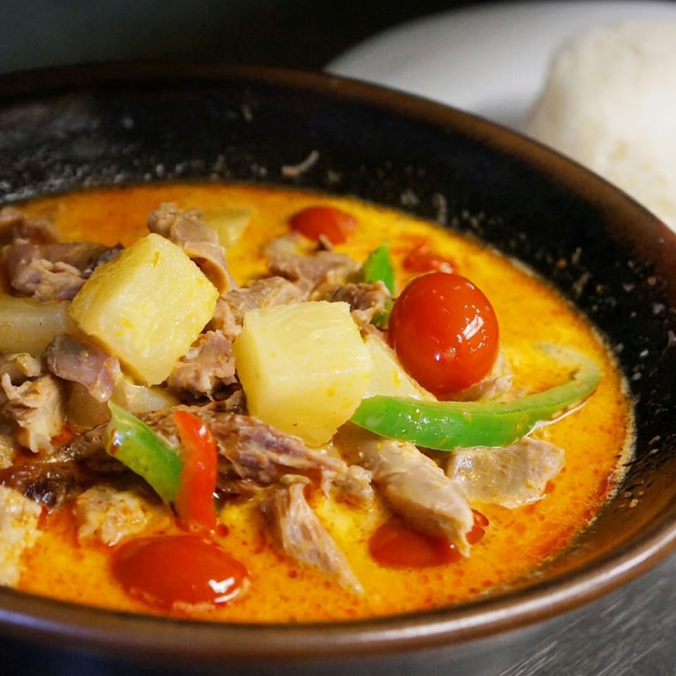 Bowl of creamy red curry with sliced meat, pineapple, bell peppers, and cherry tomatoes, served with white rice.