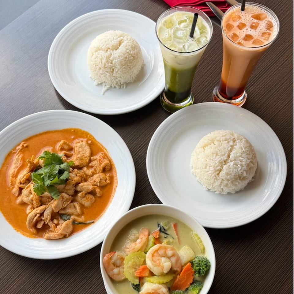 Thai meal with chicken curry, shrimp and vegetable curry, jasmine rice, and two iced drinks on a wooden table.